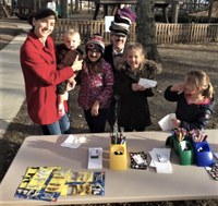 Mother and her four young daughters wearing mustaches and smiling with Ms. Liz who's dressed up as the Peddler with a stack of caps on her head