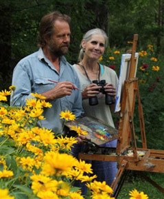 smiling couple standing together outside, trees and a canvas on an easel in the background, yellow flowers in the lower left foreground. Woman is holding binoculars and man is holding a paint pallette with paint brush